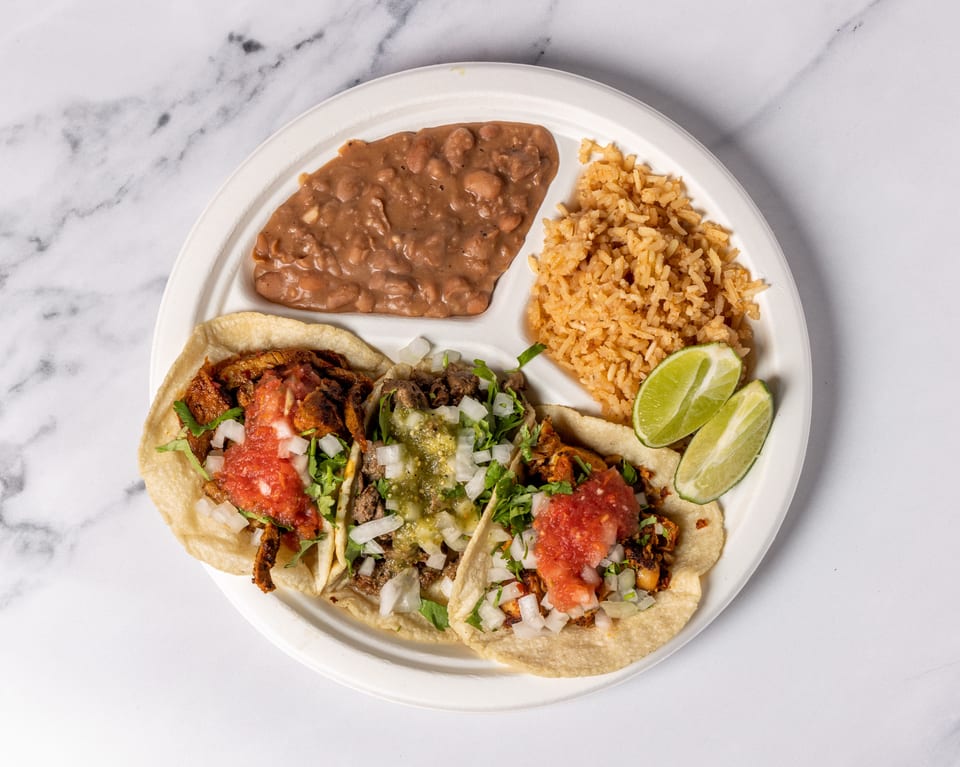 Three tacos with various toppings, rice, and beans on a white plate with lime wedges.