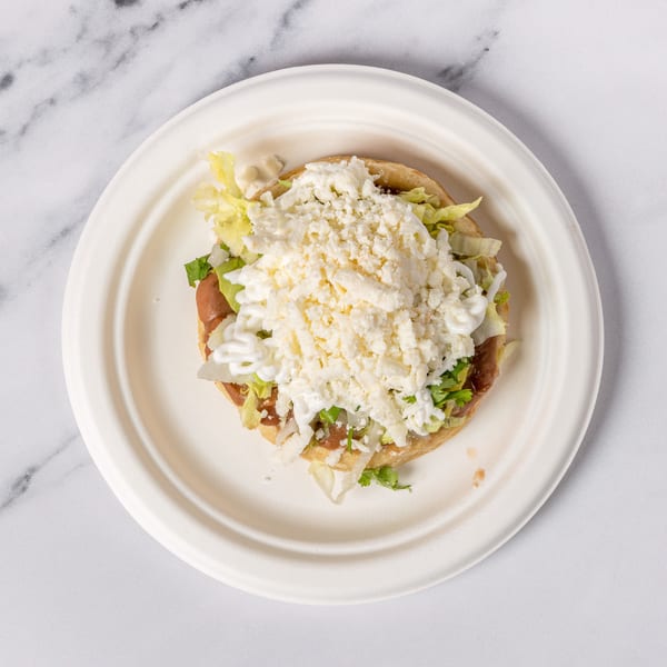 A sopes on a white plate, topped with refried beans, lettuce, sour cream, and crumbled cheese.