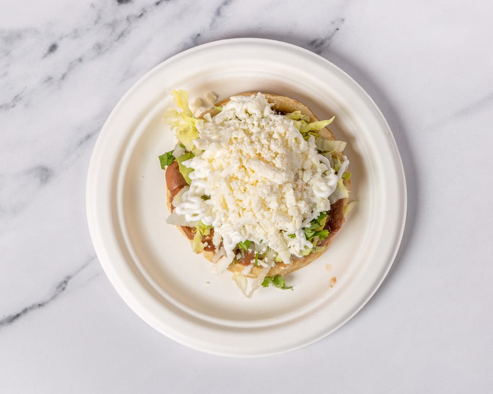 A sopes on a white plate, topped with refried beans, lettuce, sour cream, and crumbled cheese.