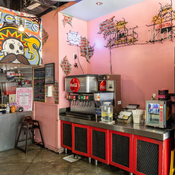Interior of a vibrant taco shop with pink walls, graffiti, neon signs, and a soda fountain.