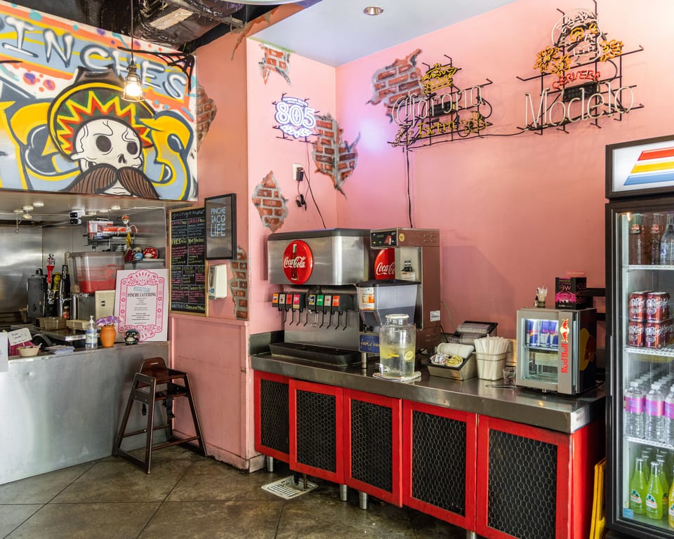 Interior of a vibrant taco shop with pink walls, graffiti, neon signs, and a soda fountain.