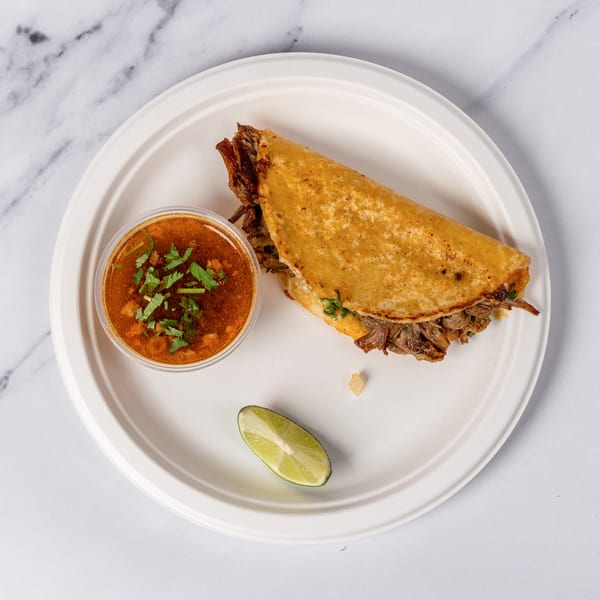 Birria taco with a side of consommé and a lime wedge on a white plate.