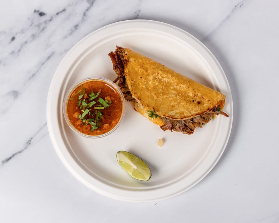 Birria taco with a side of consommé and a lime wedge on a white plate.