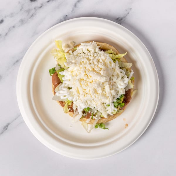 A sopes dish with refried beans, lettuce, sour cream, and crumbled white cheese on a white plate.