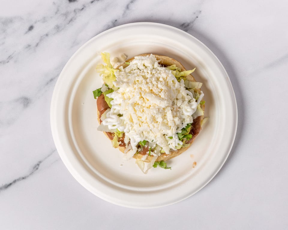 A sopes dish with refried beans, lettuce, sour cream, and crumbled white cheese on a white plate.