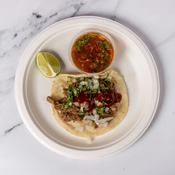A beef birria taco with onions, cilantro, and red sauce, served with consommé and a lime wedge.