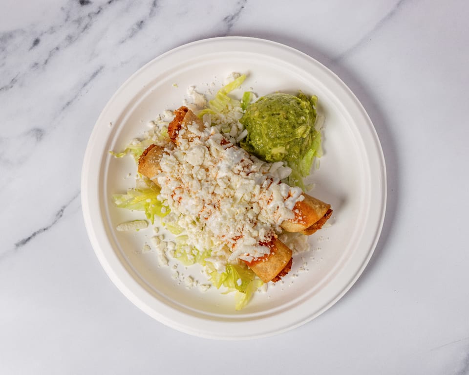 Two taquitos topped with cheese and sauce, served with guacamole and lettuce on a white plate.