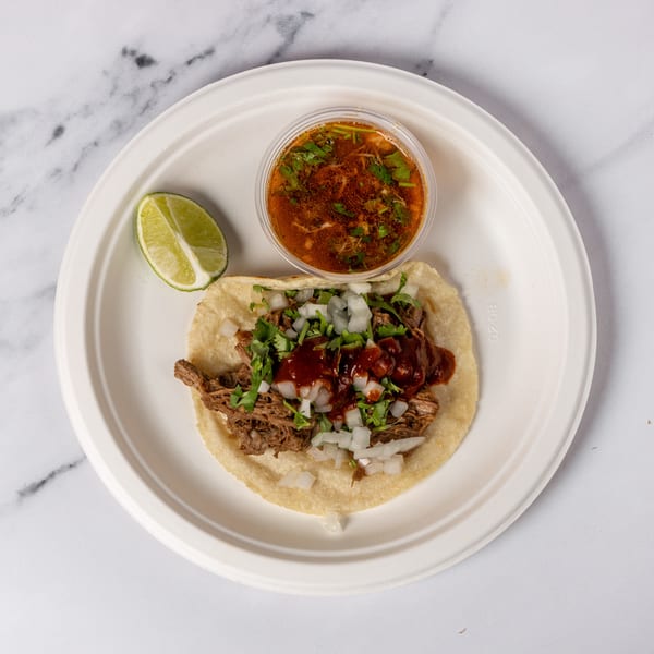 A birria taco with onions, cilantro, and red sauce, served with a side of consommé and lime.