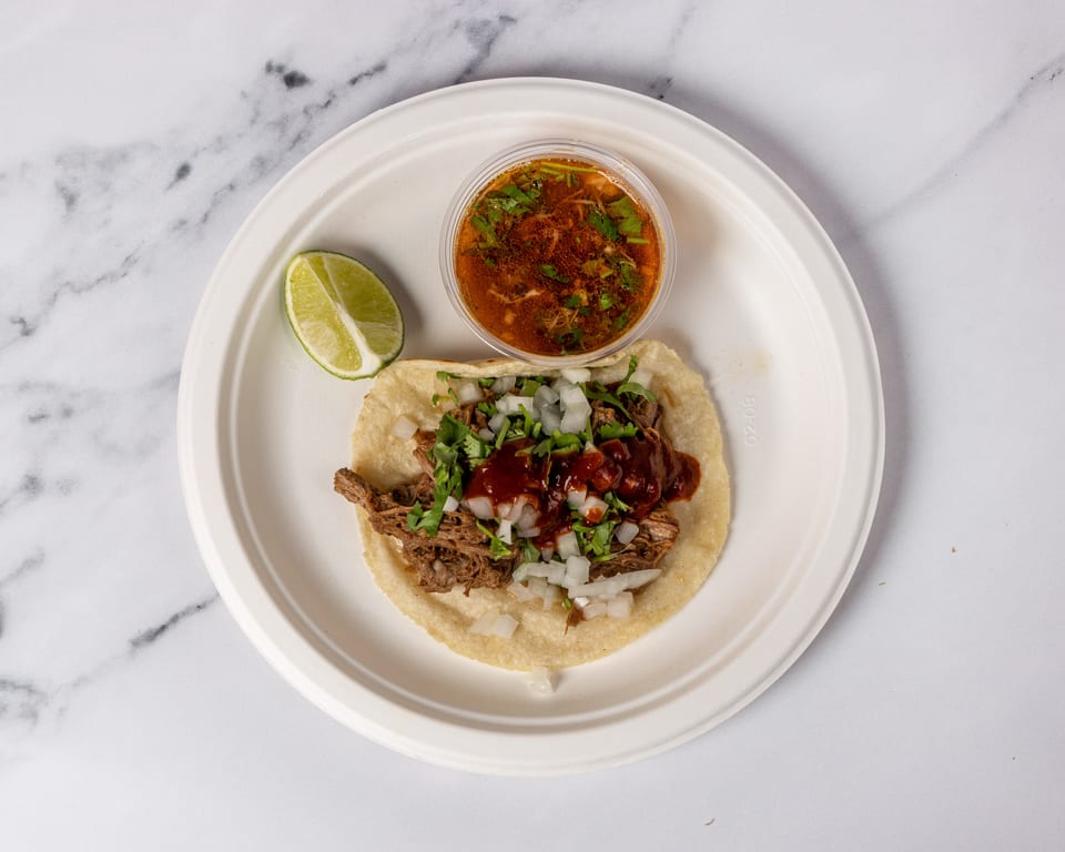 A birria taco with onions, cilantro, and red sauce, served with a side of consommé and lime.
